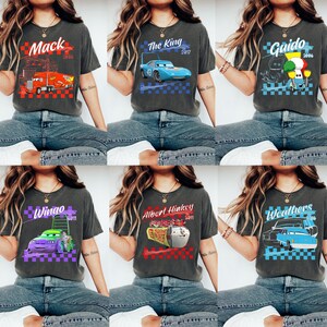 May include: Six dark gray t-shirts featuring graphics of characters from the movie Cars. Each shirt has a different character design with text, including "Mack", "The King", "Guido", "Wingo", "Albert Hinkey", and "Weathers".