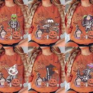 Könnte beinhalten: Orangefarbene Halloween-T-Shirts mit verschiedenen Cartoon-Charakter-Designs. Jedes Shirt zeigt einen Charakter in einem Skelettkostüm, Kürbisse, Grabsteine und das Wort "BOO!" in einem Spinnennetz-Design. Die Charaktere sind Kermit, Mater, Buzz Lightyear, Moana, Scar und Figment.
