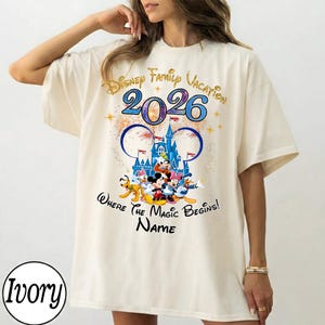 May include: Ivory-colored t-shirt featuring a colorful Disney graphic. The design includes the text "Disney Family Vacation 2026" and a castle illustration with Mickey Mouse and friends. The shirt has a relaxed fit.