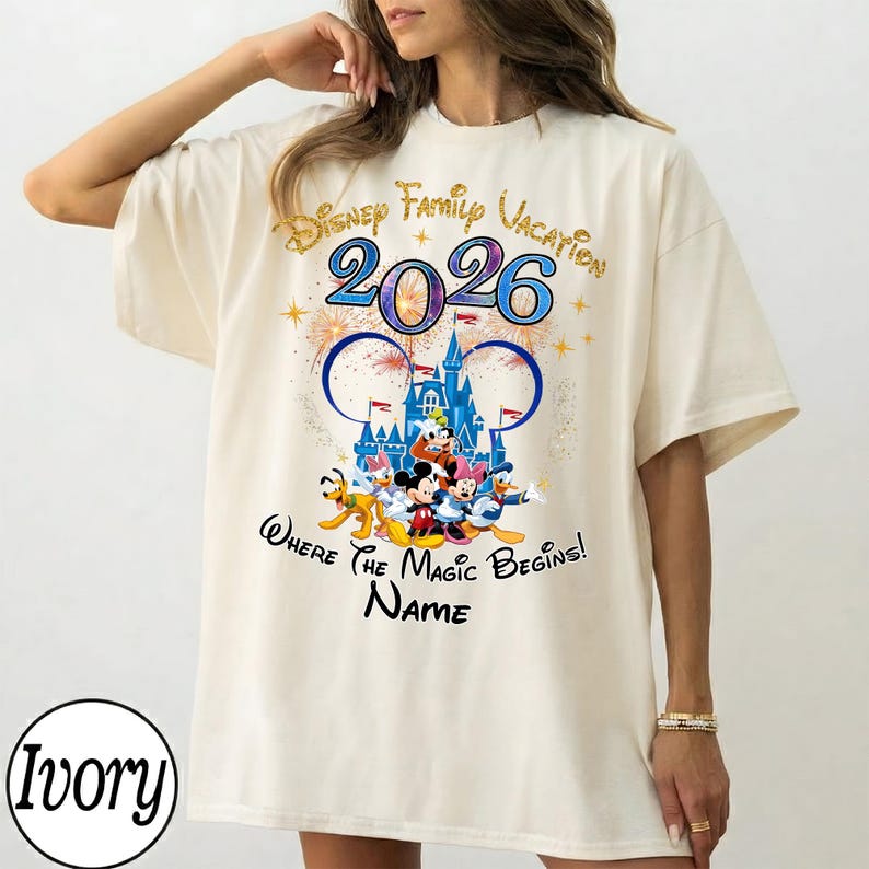 May include: Ivory-colored t-shirt featuring a colorful Disney graphic. The design includes the text "Disney Family Vacation 2026" and a castle illustration with Mickey Mouse and friends. The shirt has a relaxed fit.