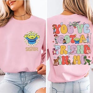 May include: A light pink sweatshirt featuring a cartoon alien with the word "SISTER" below it on the front. The back of the sweatshirt has the phrase "YOU'VE GOT A FRIEND IN ME" with various cartoon characters.