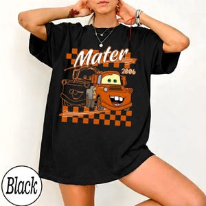 May include: Black t-shirt featuring a cartoon graphic of Mater from the movie Cars, with the text "Mater 2006" above the image. The design includes an orange and black checkered pattern.