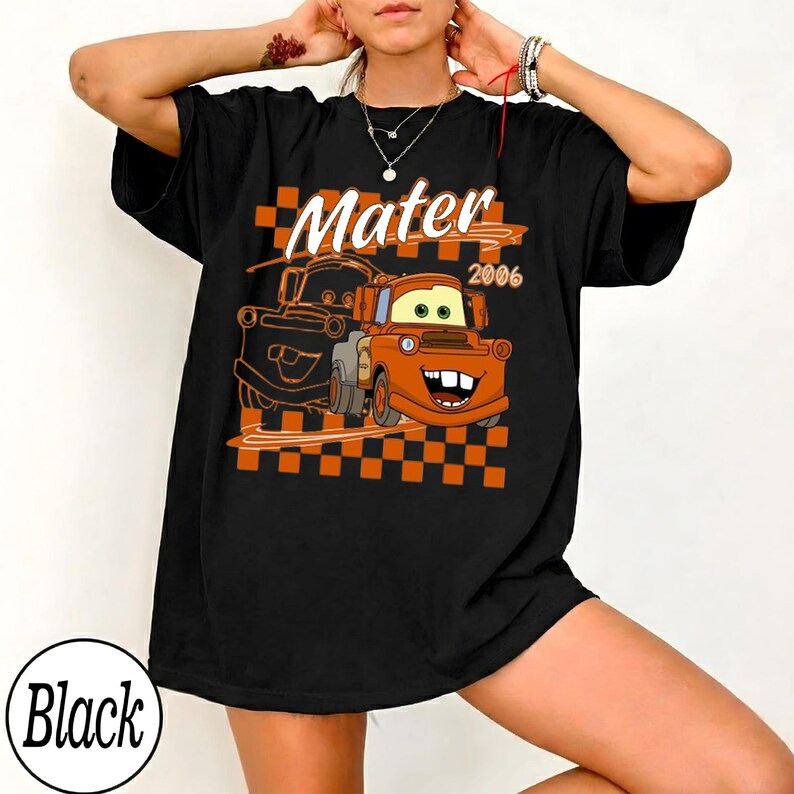 May include: Black t-shirt featuring a cartoon graphic of Mater from the movie Cars, with the text "Mater 2006" above the image. The design includes an orange and black checkered pattern.