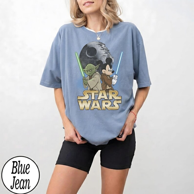 May include: A light blue t-shirt featuring a Star Wars graphic with Yoda and Mickey Mouse holding lightsabers. The Star Wars logo is in gold. The shirt is a relaxed fit.