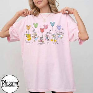 May include: A light pink t-shirt featuring a graphic of Disney characters holding Mickey Mouse-shaped balloons. The characters include Pluto, Goofy, Mickey Mouse, Minnie Mouse, Donald Duck, and Daisy Duck. The shirt has the word "Blossom" printed on the bottom left.