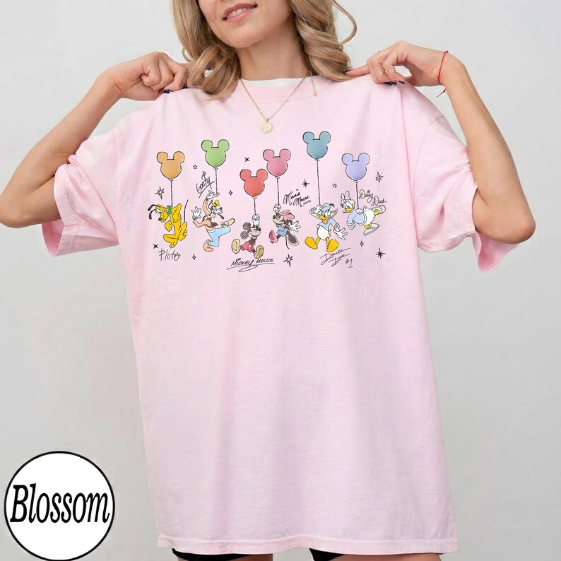 May include: A light pink t-shirt featuring a graphic of Disney characters holding Mickey Mouse-shaped balloons. The characters include Pluto, Goofy, Mickey Mouse, Minnie Mouse, Donald Duck, and Daisy Duck. The shirt has the word "Blossom" printed on the bottom left.