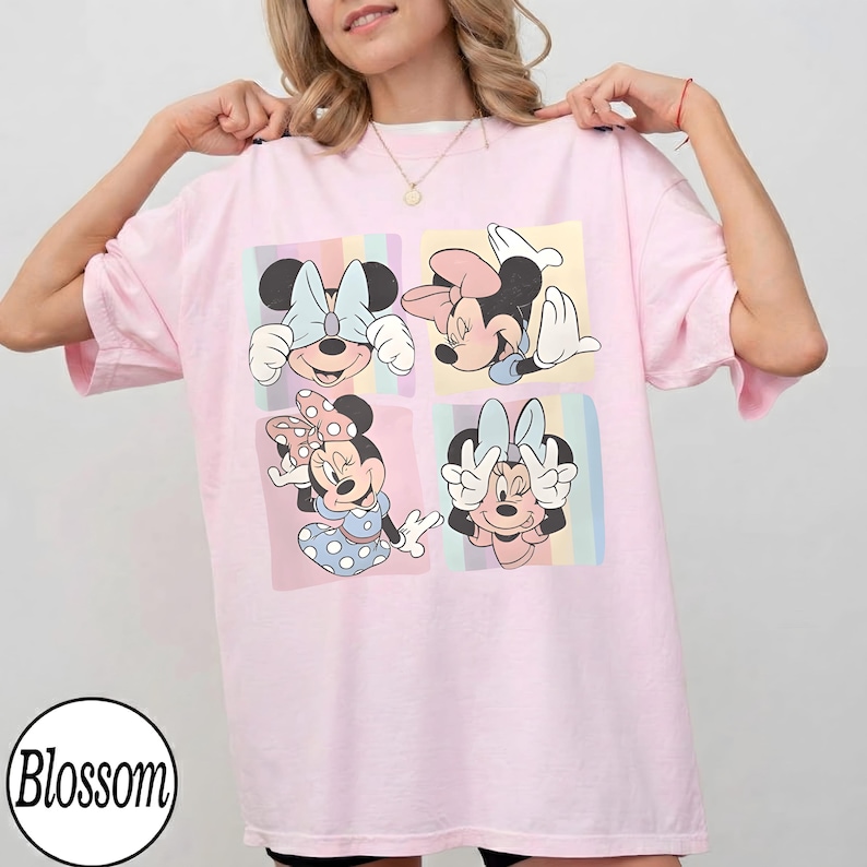 May include: A light pink t-shirt featuring a four-panel graphic of Minnie Mouse in various poses. Each panel has a pastel rainbow background. The shirt has short sleeves and a round neckline. The word "Blossom" is in a circle at the bottom left.