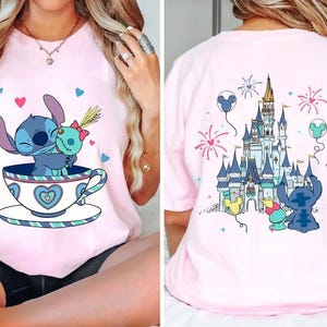 Könnte beinhalten: Hellrosa T-Shirt mit einer Cartoon-Illustration von Stitch und Angel in einer Teetasse, umgeben von Herzen. Auf der Rückseite des Shirts sind ein Schloss, Feuerwerk und Stitch mit Angel abgebildet.