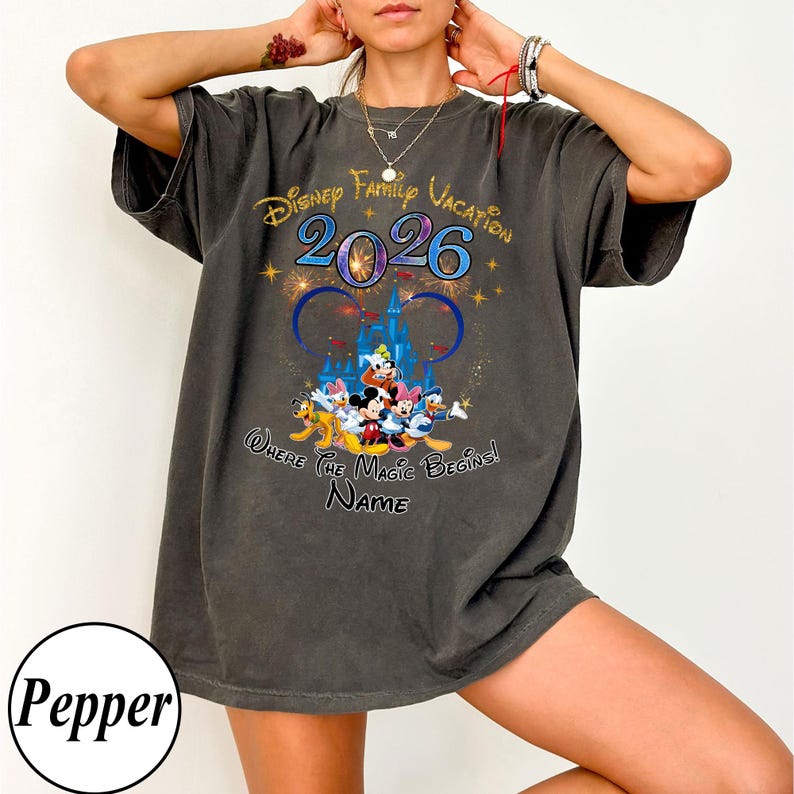 May include: Dark gray t-shirt featuring a Disney Family Vacation design for 2026. The graphic includes Disney characters, a castle, and the phrase "Where the Magic Begins!" with space for a name.