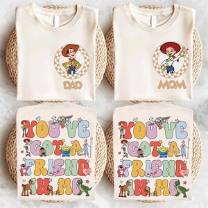 May include: Four cream-colored t-shirts. Two shirts have a Woody and Jessie cartoon design with the words "DAD" and "MOM". The other two shirts have the text "YOU'VE GOT A FRIEND IN ME" with cartoon characters.