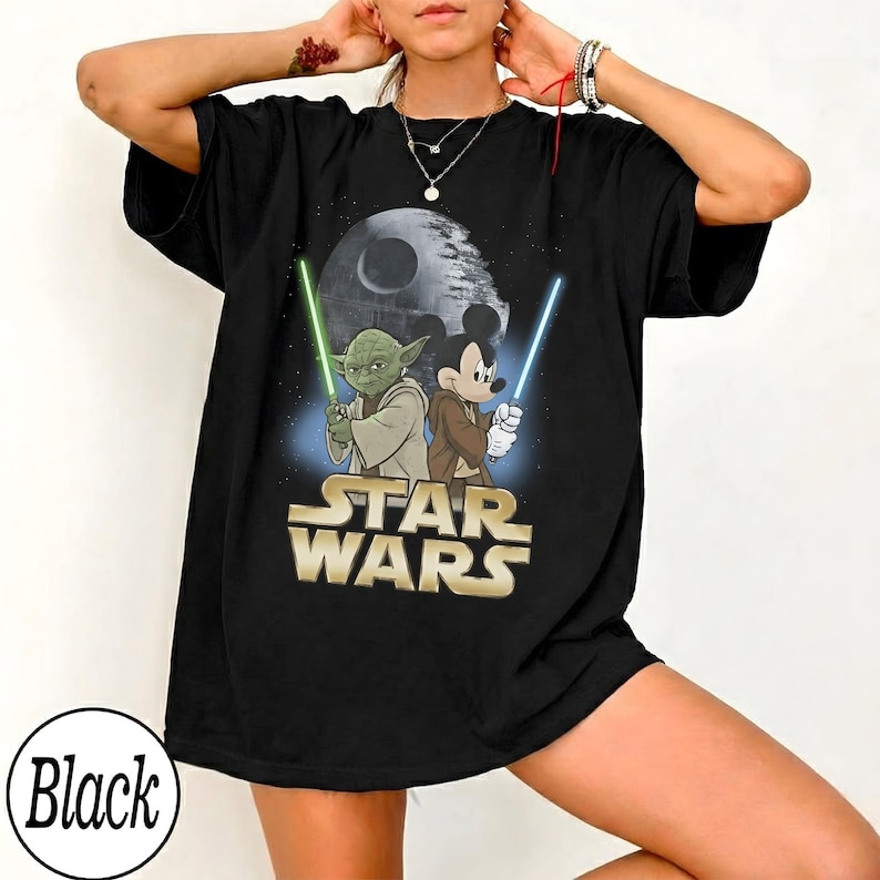 May include: Black Star Wars t-shirt featuring Yoda and Mickey Mouse holding lightsabers, with the Death Star in the background. The Star Wars logo is in gold. The shirt is black.