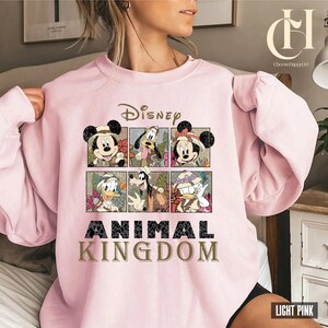 May include: Light pink sweatshirt featuring Disney characters Mickey Mouse, Minnie Mouse, Donald Duck, and Goofy. The design includes the word "Disney" above the characters and "Animal Kingdom" below. The sweatshirt is a crew neck style.