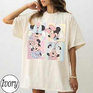 May include: Ivory-colored t-shirt featuring a four-panel design with Minnie Mouse illustrations. Each panel showcases a different pose of Minnie, set against pastel striped backgrounds. The shirt has a relaxed fit and short sleeves.