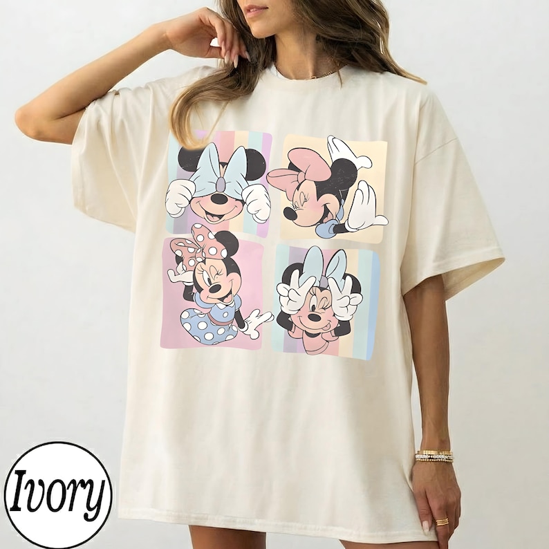 May include: Ivory-colored t-shirt featuring a four-panel design with Minnie Mouse illustrations. Each panel showcases a different pose of Minnie, set against pastel striped backgrounds. The shirt has a relaxed fit and short sleeves.