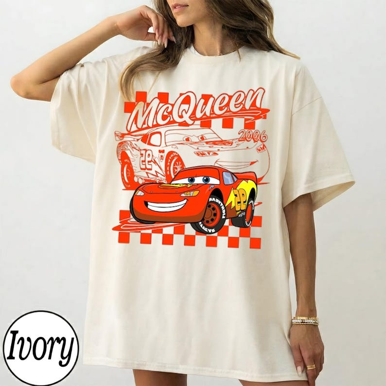 May include: Ivory-colored t-shirt featuring a graphic of Lightning McQueen from the movie Cars. The design includes the text "McQueen 2006" and a checkered pattern in red and white. The shirt has short sleeves.