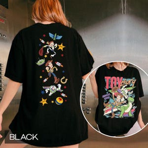 May include: A black t-shirt featuring a vertical arrangement of Toy Story characters including Woody, Buzz Lightyear, and an alien, along with stars and rockets. The front inset shows the Toy Story logo with Woody and Rex.