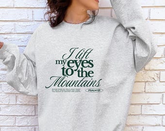 May include: A light gray sweatshirt with the text "I lift my eyes to the Mountains" in dark green script. The sweatshirt also includes the text "Psalm 121" and the phrase "Where does my help come from? My help comes from the Lord, the maker of heaven and earth."