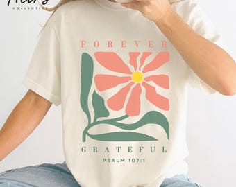 May include: A cream-colored t-shirt featuring a graphic of a large coral-pink flower with a yellow center and green leaves. The text "FOREVER" appears above the flower, and "GRATEFUL PSALM 107:1" is printed in green below the floral design.