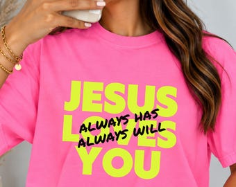 May include: A bright pink short-sleeved t-shirt featuring the text JESUS LOVES YOU in large neon yellow letters, with the words ALWAYS HAS ALWAYS WILL printed in smaller black script across the center of the design.