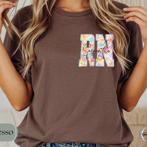 May include: Espresso-colored t-shirt with a floral design. The letters "RN" are filled with colorful flowers, and the name "Samantha" is written in script. The shirt is a casual style.