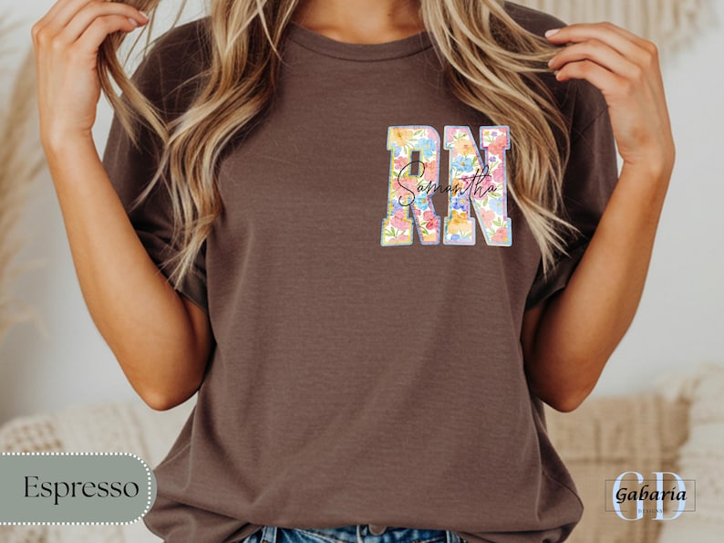 May include: Espresso-colored t-shirt with a floral design. The letters "RN" are filled with colorful flowers, and the name "Samantha" is written in script. The shirt is a casual style.
