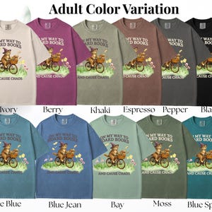 May include: A collection of short-sleeved t-shirts in various colors, each featuring a whimsical graphic of a cat riding a bicycle with books and the text "ON MY WAY TO HOARD BOOKS AND CAUSE CHAOS". Colors include ivory, berry, khaki, espresso, pepper, black, ice blue, blue jean, bay, moss, and blue spruce.