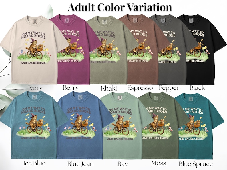May include: A collection of short-sleeved t-shirts in various colors, each featuring a whimsical graphic of a cat riding a bicycle with books and the text "ON MY WAY TO HOARD BOOKS AND CAUSE CHAOS". Colors include ivory, berry, khaki, espresso, pepper, black, ice blue, blue jean, bay, moss, and blue spruce.