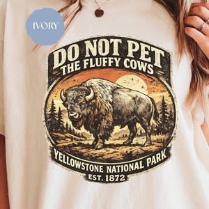 May include: Ivory-colored t-shirt featuring a vintage-style graphic. The design includes a bison illustration, the text "DO NOT PET THE FLUFFY COWS," and "Yellowstone National Park EST. 1872." The graphic has a distressed, aged look.