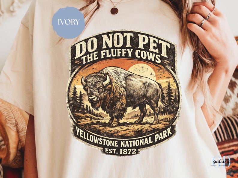 May include: Ivory-colored t-shirt featuring a vintage-style graphic. The design includes a bison illustration, the text "DO NOT PET THE FLUFFY COWS," and "Yellowstone National Park EST. 1872." The graphic has a distressed, aged look.