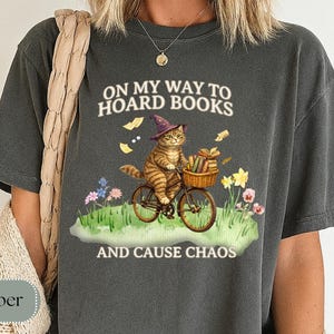 May include: Dark gray t-shirt with a whimsical graphic of a tabby cat riding a bicycle, wearing a witch's hat, and carrying books in a basket. The text reads "ON MY WAY TO HOARD BOOKS AND CAUSE CHAOS."