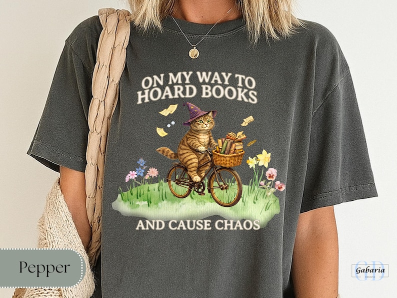 May include: Dark gray t-shirt with a whimsical graphic of a tabby cat riding a bicycle, wearing a witch's hat, and carrying books in a basket. The text reads "ON MY WAY TO HOARD BOOKS AND CAUSE CHAOS."