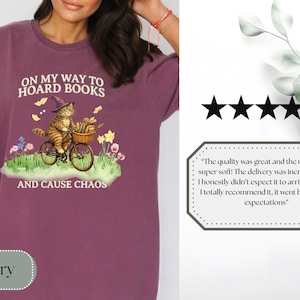 May include: Berry-colored t-shirt with a whimsical graphic of a cat in a witch hat riding a bicycle, carrying books and flowers. Text reads "ON MY WAY TO HOARD BOOKS AND CAUSE CHAOS."