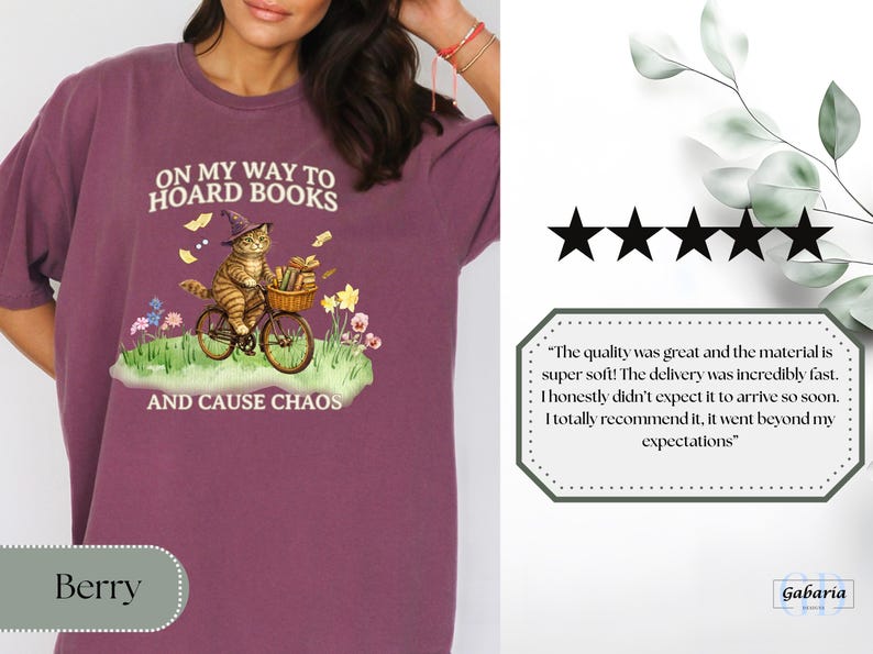 May include: Berry-colored t-shirt with a whimsical graphic of a cat in a witch hat riding a bicycle, carrying books and flowers. Text reads "ON MY WAY TO HOARD BOOKS AND CAUSE CHAOS."