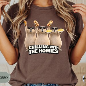 K&ouml;nnte beinhalten: Espressofarbenes T-Shirt mit einer Grafik von drei Capybaras, die Sonnenbrillen tragen und Hot Dogs und Teller mit Essen halten. Der Text "CHILLING WITH THE HOMIES" befindet sich darunter. Das Shirt ist einfarbig.