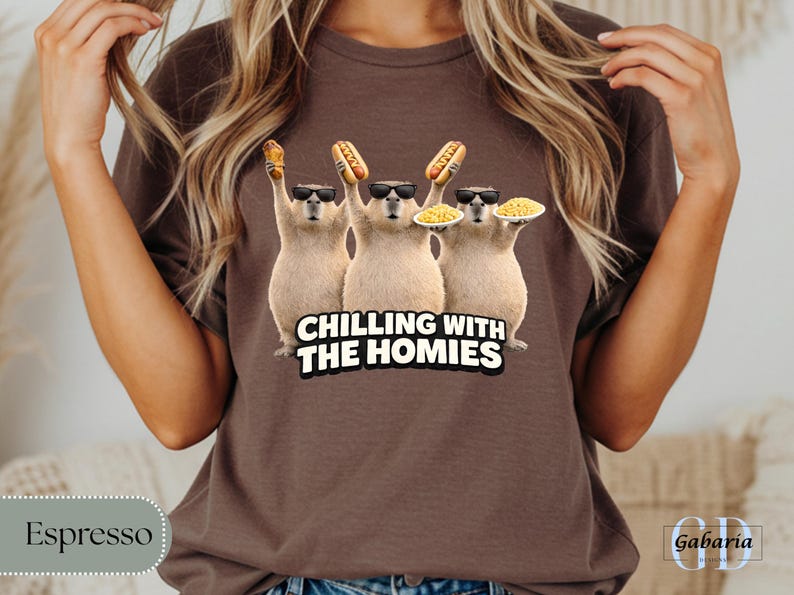 K&ouml;nnte beinhalten: Espressofarbenes T-Shirt mit einer Grafik von drei Capybaras, die Sonnenbrillen tragen und Hot Dogs und Teller mit Essen halten. Der Text "CHILLING WITH THE HOMIES" befindet sich darunter. Das Shirt ist einfarbig.
