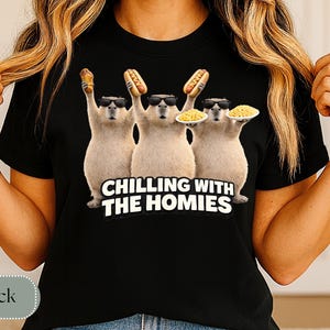 K&ouml;nnte beinhalten: Schwarzes T-Shirt mit einer Grafik von drei Capybaras, die Sonnenbrillen tragen und Essen halten. Der Text "CHILLING WITH THE HOMIES" ist darunter aufgedruckt. Die Capybaras halten einen Hotdog, einen Maiskolben und einen Teller Pasta.