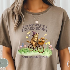 May include: Khaki-colored t-shirt featuring a whimsical illustration of a cat riding a bicycle with a basket of books. The cat wears a wizard hat, and the text reads "ON MY WAY TO HOARD BOOKS AND CAUSE CHAOS."