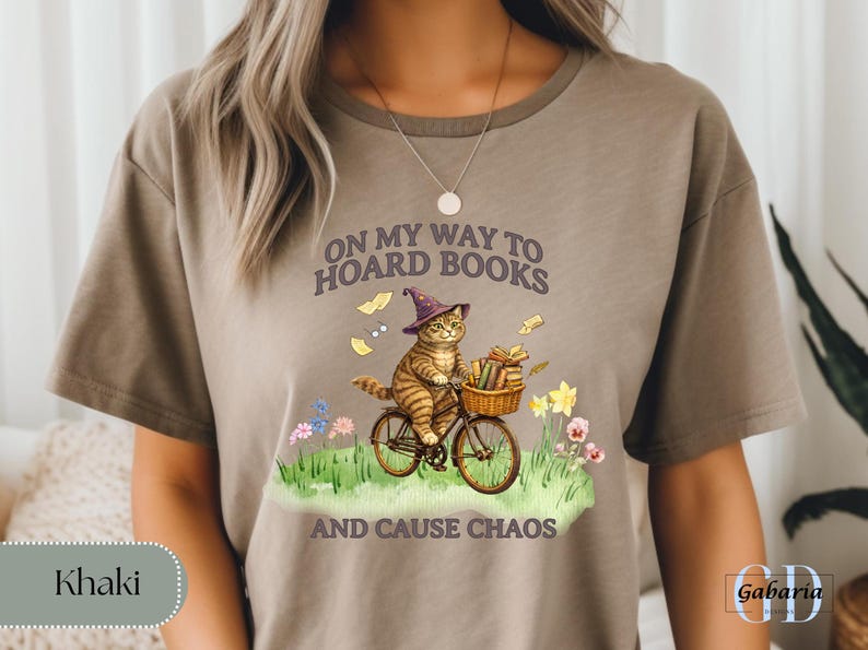 May include: Khaki-colored t-shirt featuring a whimsical illustration of a cat riding a bicycle with a basket of books. The cat wears a wizard hat, and the text reads "ON MY WAY TO HOARD BOOKS AND CAUSE CHAOS."