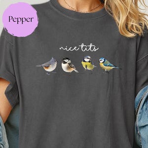 May include: Dark gray t-shirt with the text "nice tits" above a watercolor illustration of four small birds. The shirt has a relaxed fit and is made of a soft material. A purple circle with the word "Pepper" is in the upper left corner.