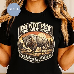 May include: Black t-shirt featuring a vintage-style graphic of a bison in Yellowstone National Park. The design includes the text "DO NOT PET THE FLUFFY COWS" and "Yellowstone National Park EST. 1872". The shirt has a crew neck and short sleeves.