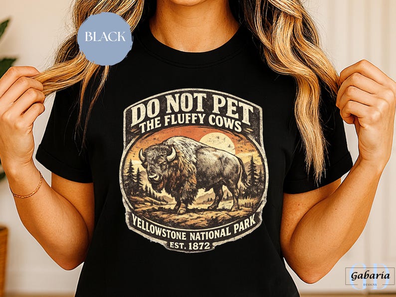 May include: Black t-shirt featuring a vintage-style graphic of a bison in Yellowstone National Park. The design includes the text "DO NOT PET THE FLUFFY COWS" and "Yellowstone National Park EST. 1872". The shirt has a crew neck and short sleeves.