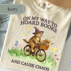 May include: Ivory-colored t-shirt with a whimsical graphic of a cat riding a bicycle, wearing a wizard hat, and carrying books in a basket. The text reads "ON MY WAY TO HOARD BOOKS AND CAUSE CHAOS."