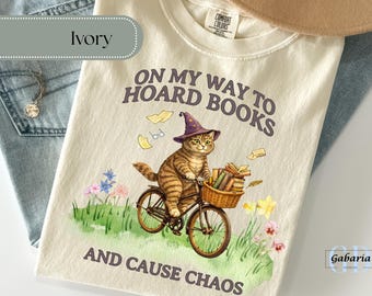May include: Cream-coloured t-shirt featuring a cat on a bicycle, wearing a wizard's hat, and carrying books. The text says "ON MY WAY TO HOARD BOOKS AND CAUSE CHAOS". The shirt is folded on denim.