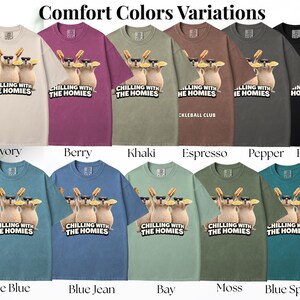 K&ouml;nnte beinhalten: Eine Auswahl an Comfort Colors T-Shirts in verschiedenen Farben, darunter Elfenbein, Beere, Khaki und Schwarz. Jedes Shirt zeigt eine Grafik von drei Zeichentrickkatzen mit Sonnenbrillen und Hotdogs, mit dem Text "CHILLING WITH THE HOMIES".