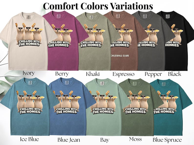 K&ouml;nnte beinhalten: Eine Auswahl an Comfort Colors T-Shirts in verschiedenen Farben, darunter Elfenbein, Beere, Khaki und Schwarz. Jedes Shirt zeigt eine Grafik von drei Zeichentrickkatzen mit Sonnenbrillen und Hotdogs, mit dem Text "CHILLING WITH THE HOMIES".