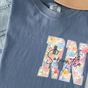 May include: A blue Comfort Colors t-shirt with a floral design. The letters "RN" are filled with colorful flowers and the name "Samantha" is written in cursive script across the letters. A gold necklace and jeans are visible.