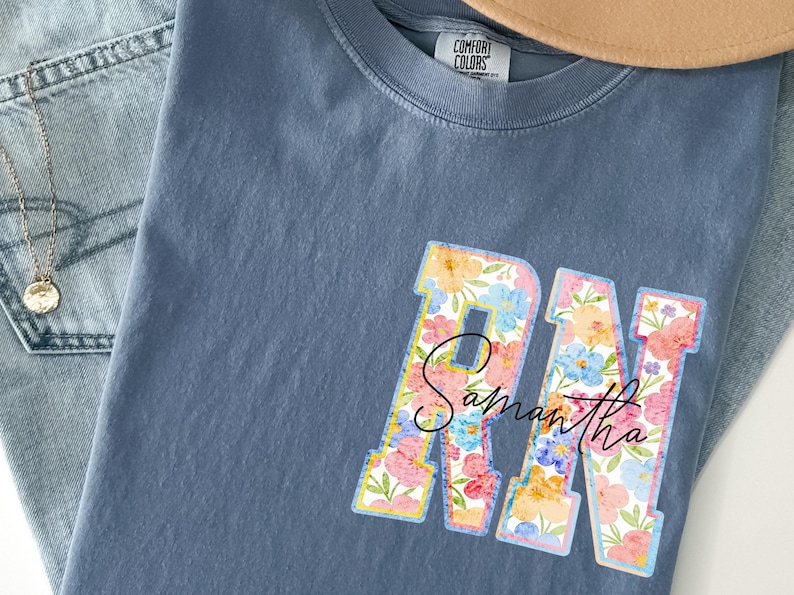 May include: A blue Comfort Colors t-shirt with a floral design. The letters "RN" are filled with colorful flowers and the name "Samantha" is written in cursive script across the letters. A gold necklace and jeans are visible.