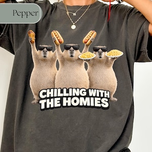 K&ouml;nnte beinhalten: Dunkelgraues T-Shirt mit einer Grafik von drei Capybaras mit Sonnenbrillen, die Hotdogs und Teller mit Essen halten. Der Text "CHILLING WITH THE HOMIES" ist unter den Capybaras in Wei&szlig; und Schwarz gedruckt.