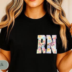 May include: Black t-shirt featuring the letters "RN" in a floral design, with the name "Samantha" in script below. The shirt is a crew neck style.