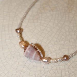 May include: A delicate necklace featuring a natural seashell pendant with pink and white stripes. The necklace is adorned with small, lustrous pearls in shades of pink and white, creating an elegant and beachy aesthetic.